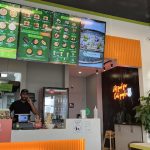 A modern restaurant with a green menu board, a neon sign, and a staff member at the counter.